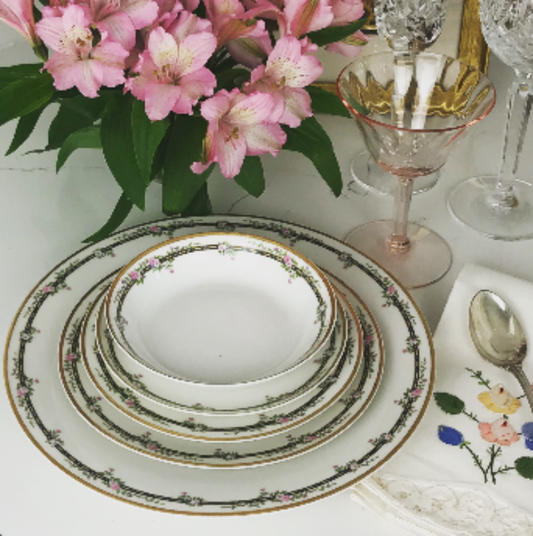 A set of white china dishes with a gold rim, dark border and pink floral details. This image features a stack starting with a large dinner plate or charger, a smaller salad plate, then a bread plate on top of which there is a large soup bowl and a smaller fruit bowl. In the background there are pink and crystal glasses and a floral arrangement.